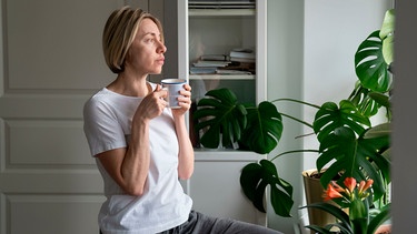 Eine Frau trinkt Kaffee aus einer Tasse und sieht aus dem Fenster | Bild: mauritius images / Dmitry Marchenko / Alamy / Alamy Stock Fotos