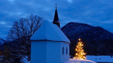 Kapelle im Winter mit einem leuchtenden Weihnachtsbaum davor. | Bild: mauritius images / Westend61 / Lisa und Wilfried Bahnmüller