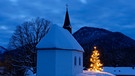 Kapelle im Winter mit einem leuchtenden Weihnachtsbaum davor. | Bild: mauritius images / Westend61 / Lisa und Wilfried Bahnmüller