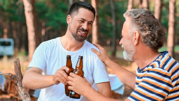 Ein jüngerer und ein älterer Mann sitzen in einem Kiefernwald und trinken Bier aus der Flasche | Bild: mauritius images / Pixel-shot / Alamy / Alamy Stock Photos