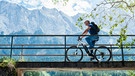 Mann ist auf seinem E-Bike unterwegs und fährt über eine Brücke. Im Hintergrund Berge. | Bild: mauritius images / Westend61 / Daniel Ingold