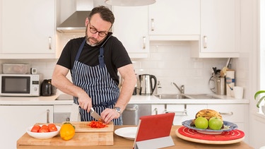 Ein Mann schneidet in einer hellen Küche Gemüse auf einem Brett, telefoniert dabei und hat ein Rezept auf einem Tablet aufgerufen | Bild: mauritius images / Westend61 / Junge