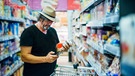 Mann hat ein Glas Nuss-Nougat-Creme in einem Supermarkt in der Hand und sieht auf sein Smartphone | Bild: mauritius images / Westend61 / Jesús Martinez