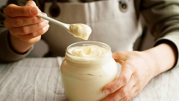 Frau hält Löffel mit Mayonnaise in der Hand, vor sich ein Mayonnaise-Glas | Bild: mauritius images / Tutatama / Alamy / Alamy Stock Photos