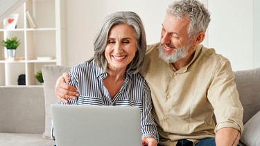 Ein fröhliches Paar mittleren Alters sitzt auf einer Couch mit einem Laptop und shoppt online | Bild: mauritius Bilder / Insta_photos / Alamy / Alamy Stock Fotos