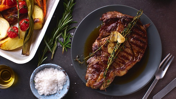 Auf einem dunklen Teller liegt ein Ribeye Angus Prime Steak, garniert mit Kräutern und Knoblauch. | Bild: mauritius images / The Picture Pantry / Anastasia Nurullina