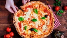 Auf einem Holztisch steht ein Teller mit einer großen Pizza Margherita. Eine Hand zieht ein Stück davon vom Teller. | Bild: mauritius images / Haris Calkic / Alamy / Alamy Stock Photos