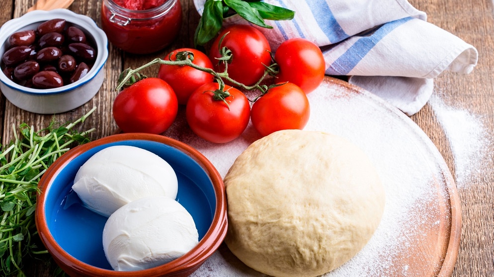 Hefeteig und Pizzazutaten wie Oliven und Tomaten liegen auf einem Holzbrett | Bild: mauritius images / Istetiana / Alamy / Alamy Stock Photos