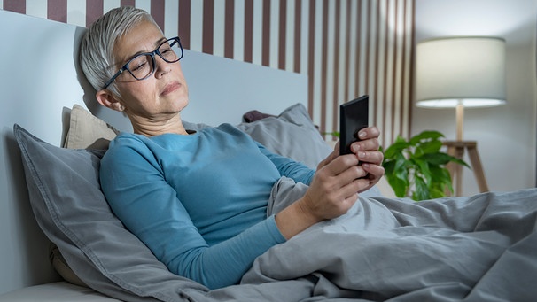 Frau liegt im Bett und schaut kritisch auf ihr Smartphone | Bild: mauritius images / Science Photo Library / Microgen Images