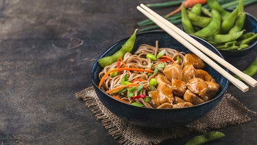 Asiatische Nudeln mit Huhn, Gemüse in einer Schüssel, rustikaler Holzhintergrund. Soba-Nudeln, Teriyaki-Huhn, Edamame, Essstäbchen | Bild: mauritius images / Olena Yeromenko / imageBROKER