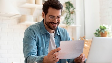 Mann liest an einem Schreibtisch zu Hause einen Brief und freut sich | Bild: mauritius images / Aleksandr Davydov / Alamy / Alamy Stock Photos