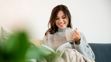 Frau trinkt eine Tasse Tee auf einem Sofa. | Bild: mauritius images / Westend61 RF / Robijn Page