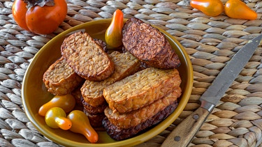 Tempeh, frittiert, auf geflochtenem Korb, dazu Flaschentomaten und eine Kaki | Bild: mauritius images / foodcollection / Dr. Martin Baumgärtner