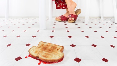 Heruntergefallener Marmeladentoast liegt auf dem Boden in einer Küche, dahinter die Füße einer Frau | Bild: mauritius images / Thomas Schultze / Volume3