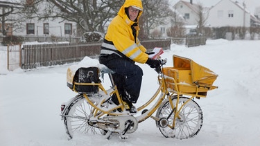 Postzusteller steht mit seinem Fahrrad im Schnee. | Bild: mauritius images / Josef Schafnitzel / imageBROKER