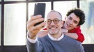 Ein Vater macht auf einer Treppe vor einem Fenster ein Selfie mit seinem jugendlichen Sohn. | Bild: mauritius images / Cavan Images