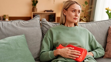 Eine Frau sitzt mit einer roten Wärmflasche auf dem Bauch auf dem Sofa | Bild: mauritius images / SeventyFour Images / Alamy / Alamy Stock Photos