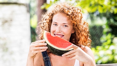 Eine Frau hält ein Stück Wassermelone in den Händen und lächelt. | Bild: mauritius images / Westend61 / Jo Kirchherr