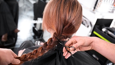 Hand einer Friseurin schneidet den geflochtetenen Zopf einer Frau ab. | Bild: mauritius images / Pixel-shot / Alamy / Alamy Stock Photos