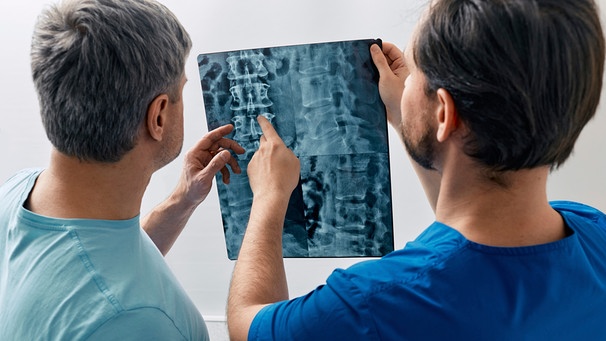 Zwei Männer, ein Patient und ein Mediziner, sehen sich die Röntgenaufnahme einer Wirbelsäule an | Bild: mauritius images / Fotothek der Wissenschaft / Peakstock