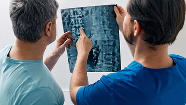 Zwei Männer, ein Patient und ein Mediziner, sehen sich die Röntgenaufnahme einer Wirbelsäule an | Bild: mauritius images / Fotothek der Wissenschaft / Peakstock