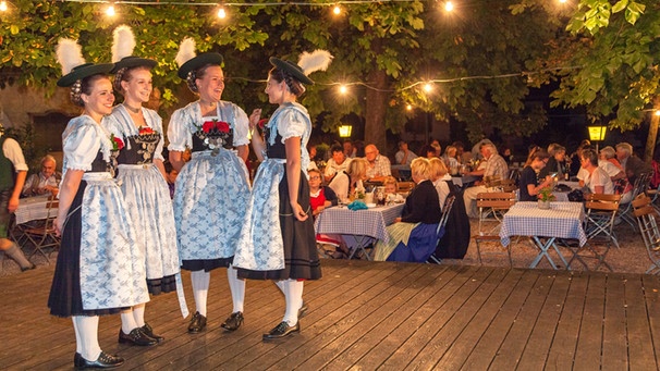 Vier junge Frauen in Tracht stehen auf einer Bühne und unterhalten sich. | Bild: mauritius images / Christian Bäck