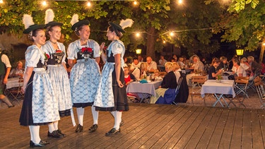 Vier junge Frauen in Tracht stehen auf einer Bühne und unterhalten sich. | Bild: mauritius images / Christian Bäck