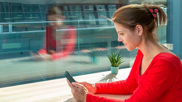 Eine Frau sitzt an einem Fenster und hat ein Smartphone in der Hand | Bild: mauritius images / Bohemama / Alamy / Alamy Stock Photos