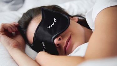 Frau liegt in einem Bett und schläft mit einer Schlafmaske | Bild: mauritius images / Westend61 / gpointstudio