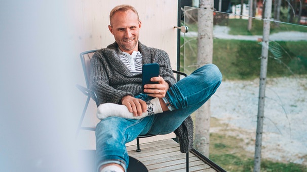 Mann entspannt mit seinem Handy auf dem Balkon | Bild: mauritius images / Creativephototeam / Alamy / Alamy Stock Photos