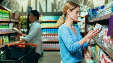 Eine Frau hält in einem Supermarkt eine Packung in der Hand und prüft die Angaben | Bild: mauritius images / Alamy Stock Photos / Liubomyr Vorona