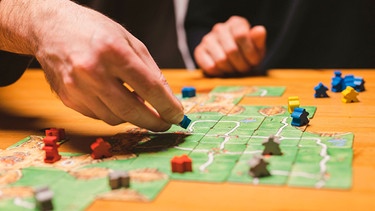 Hand über dem Brettspiel "Carcassonne" mit einer Spielfigur | Bild: mauritius images / Ksenia Shestakova / Alamy / Alamy Stock Photos