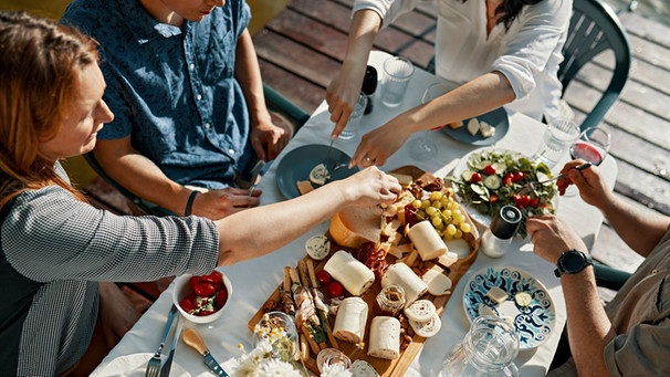 Freunde sitzen zusammen und essen Käse an einem Tisch an einem See | Bild: mauritius images / Westend61 / Zeljko Dangubic