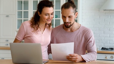 Ein Paar steht in der Küche an einem Laptop und liest einen Brief | Bild: mauritius images / Aleksandr Davydov / Alamy / Alamy Stock Photos