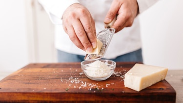 Parmesan wird in einer Küche von einem Mann in Kochjacke gerieben | Bild: mauritius images / Westend61 / Josep Rovirosa