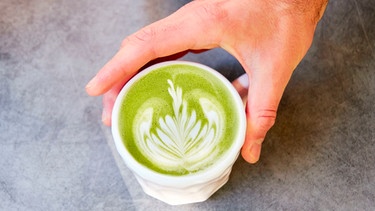 Hand reicht Tasse mit einem Matchateegetränk über den Tresen | Bild: mauritius images / Iryna Pospikh / Alamy / Alamy Stock Photos