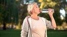 Eine Frau trinkt Vitamin-Wasser aus einer Flasche. Sie ist im Park und die Sonne scheint auf sie. | Bild: mauritius images / Ievgen Chabanov / Alamy / Alamy Stock Photos