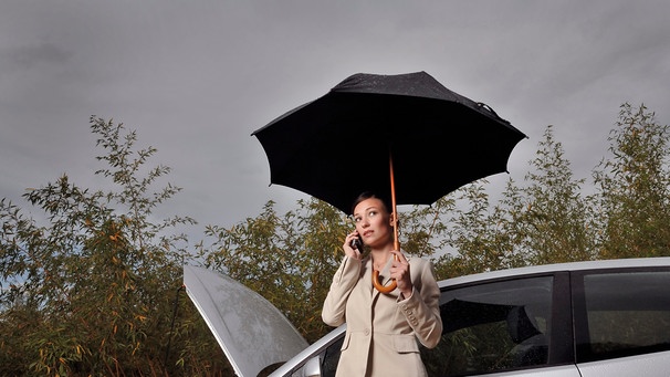 Frau telefoniert während eines Gewitters vor ihrem defekten Auto mit Regenschirm in der Hand | Bild: picture alliance  Cultura  Luc Beziat