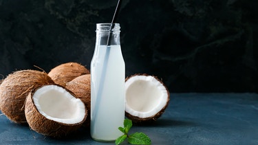 Kokoswasser in einer Glasflasche mit Strohalm neben aufgeschnittenen Kokosnüssen | Bild: mauritius images / Pixel-shot / Alamy / Alamy Stock Photos