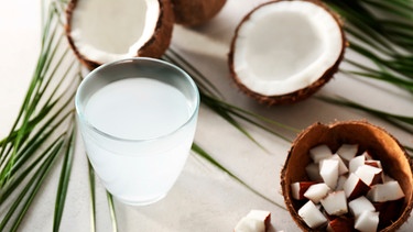 Ein Glas mit Kokoswasser neben Palmblättern und aufgeschnittenen Kokosnüssen | Bild: mauritius images / Pixel-shot / Alamy / Alamy Stock Photos