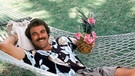 Tom Selleck in der Rolle des Privatdetektives "Magnum" in einer Hängematte mit Ananas-Drink. | Bild: picture-alliance/dpa