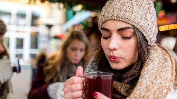 Eine junge Frau hält einen Glühwein in beiden Händen und pustet ihn kühl. | Bild: mauritius images / Sebastian Siebert Siebert / Alamy / Alamy Stock Fotos