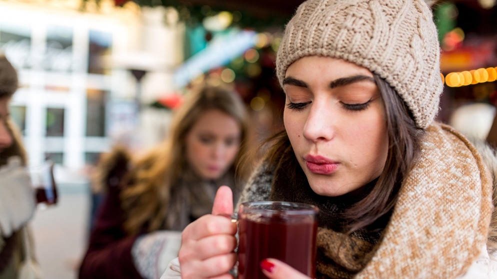 Eine junge Frau hält einen Glühwein in beiden Händen und pustet ihn kühl. | Bild: mauritius images / Sebastian Siebert Siebert / Alamy / Alamy Stock Fotos