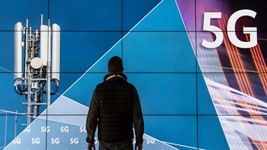 ARCHIV - 18.03.2019, Rheinland-Pfalz, Mainz: Ein Mann steht im Gebäude der Bundesnetzagentur vor einer Leinwand mit der Aufschrift «5G». Sofern das Land Rheinland-Pfalz eine von fünf geplanten Modellregionen für das neue Mobilfunknetz der fünften Generation (5G) bekommt, soll die nach dem Willen des Landes in  Kaiserslautern sein. Foto: Boris Roessler/dpa +++ dpa-Bildfunk +++ | Bild: dpa-Bildfunk/Boris Roessler