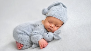Ein Baby schläft mit einem Kuscheltier im Arm auf einer grauen Decke.
| Bild: stock.adobe.com/Oksana Vashchuk