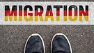 Schuhe von einem Mann auf dem Asphalt mit dem Schriftzug in Deutschland Nationalfarben: Migration | Bild: picture alliance / CHROMORANGE | CHROMORANGE