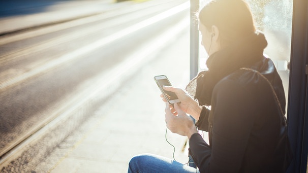 Eine Frau wartet an der Haltestelle und hört einen Podcast über das Smartphone | Bild: BR