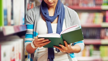 Eine Frau mit einem Buch in der Hand steht vor einem Bücherregal | Bild: colourbox.com