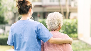 Rückenansicht einer Pflegekraft und einer älteren Frau in einem Garten.
| Bild: stock.adobe.com/interstid