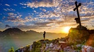 Bergsteigerin steht bei Sonnenuntergang am Gipfelkreuz der Brettgabel, hinten Watzmann, Hochkalter und Reiteralpe, Berchtesgadener Alpen, Nationalpark Berchtesgaden, Schönau am Königssee, Berchtesgadener Land, Bayern, Deutschland, Europa | Bild: picture alliance / imageBROKER | Herbert Berger